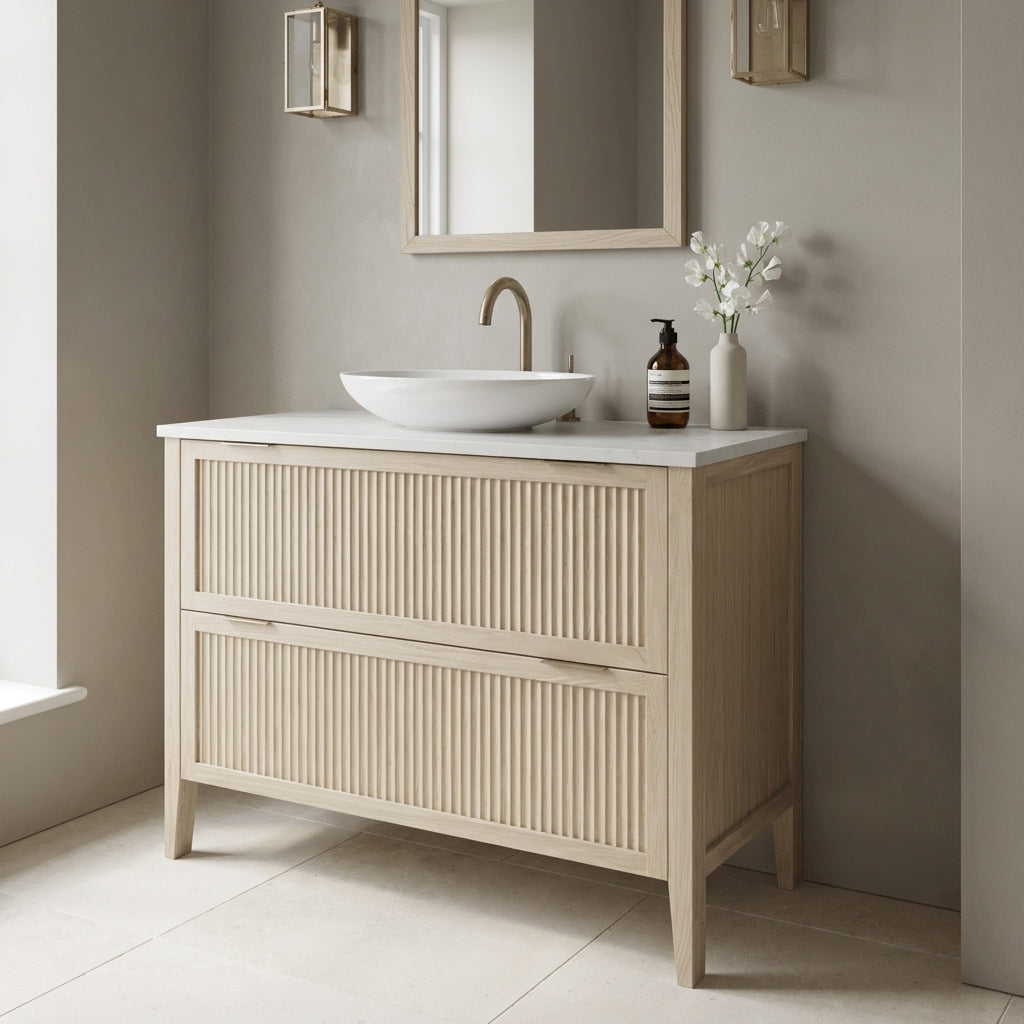 Mockridge Design Clifton 1000mm floor-standing single vanity in white stained oak with soft cream-blonde limed character and prominent vertical grain figuring across vertical fluted drawer fronts set within shaker frame borders, Calacatta quartz worktop, oval ceramic sit-on basin and brushed aged brass deck-mounted vessel mixer tap, styled in a tonally confident English bathroom with mid-greige painted chalky plaster walls and honed pale cream limestone floor