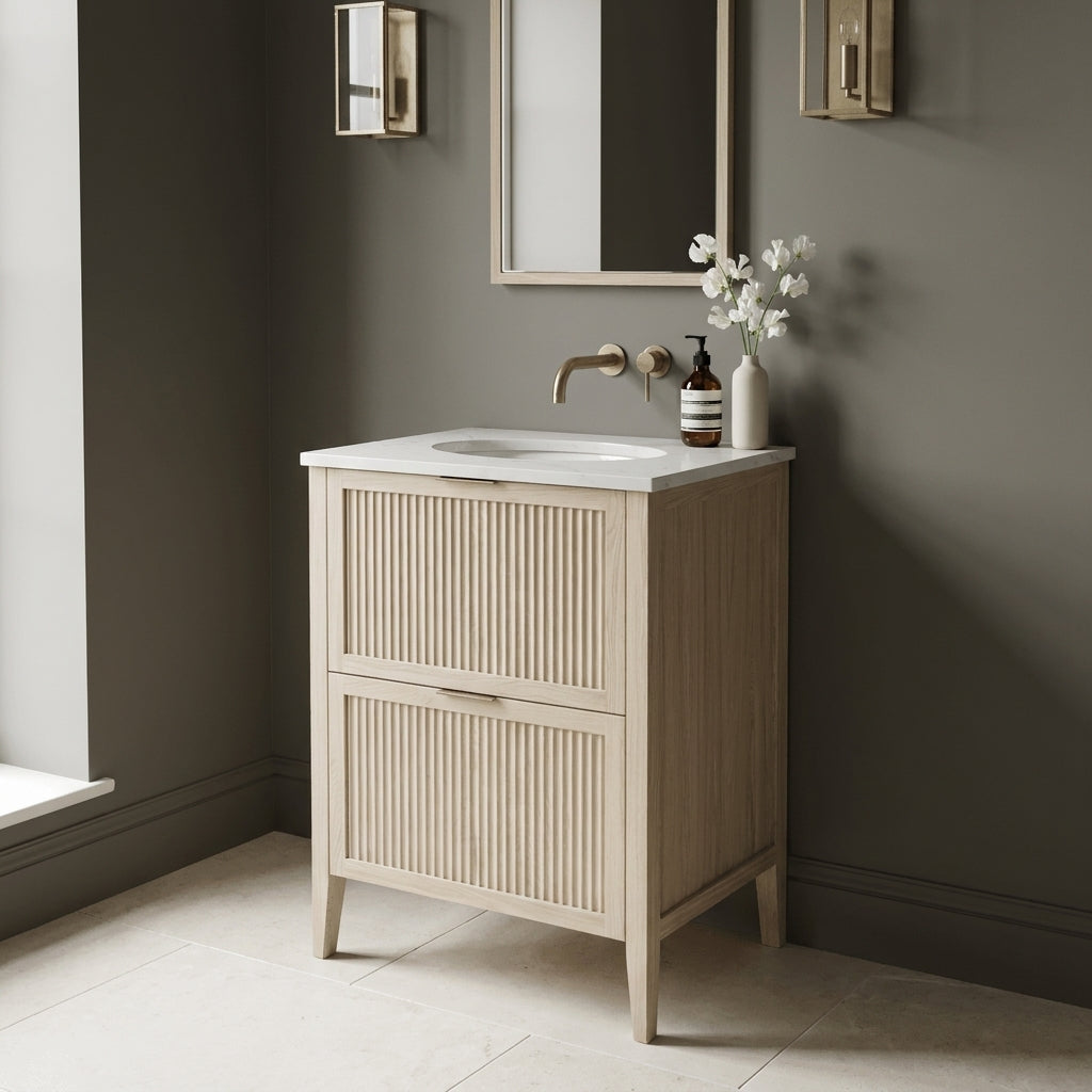 Mockridge Design Clifton 600mm floor-standing single undermount vanity in white stained oak with soft cream-blonde limed character and prominent vertical grain figuring across vertical fluted drawer fronts set within shaker frame borders, Calacatta quartz worktop with integrated undermount basin and brushed aged brass wall-mounted mixer tap, styled in a tonally confident English bathroom with mid-greige painted chalky plaster walls and honed pale cream limestone floor