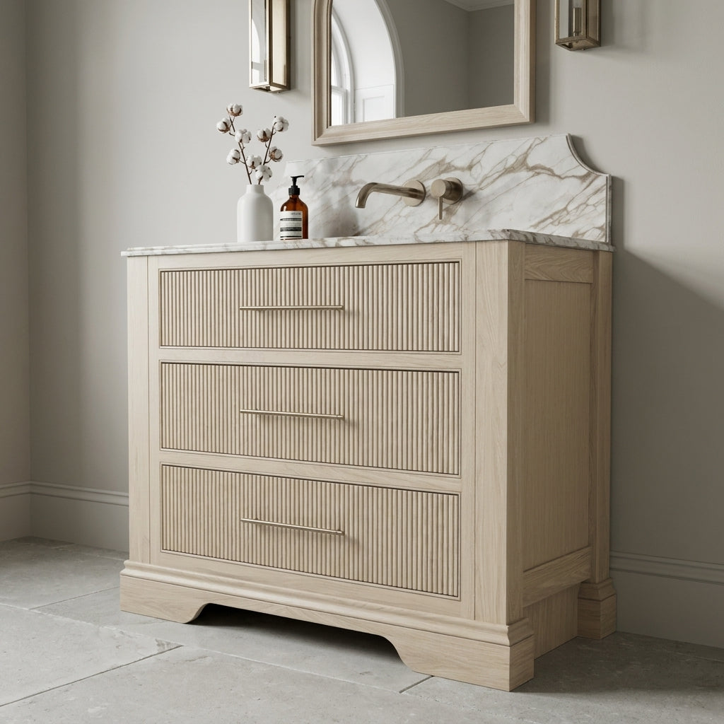 Mockridge Design Kingsbridge 1000mm floor-standing single vanity in White stained oak with very pale temperature-neutral character and visible vertical grain and reeded drawer fronts, dramatic shaped Calacatta marble backsplash, oval undermount basin and warm brushed brass wall-mounted mixer tap, styled in a deluxe grand traditional English bathroom with Farrow and Ball Purbeck Stone painted walls and honed Ancaster stone limestone floor