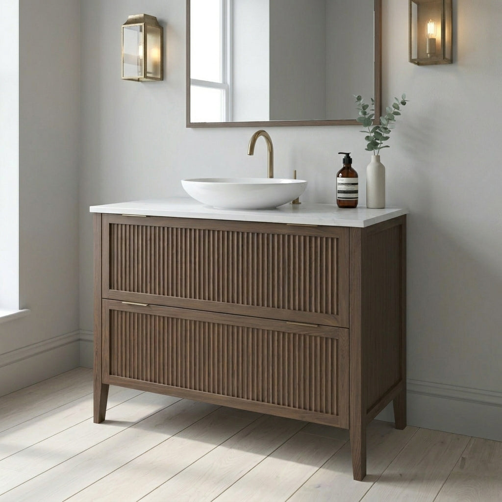 Mockridge Design Clifton 1000mm floor-standing single vanity in chocolate stained oak with smoky mid-brown grain figuring and vertical fluted drawer fronts set within shaker frame borders, Calacatta quartz worktop, oval ceramic sit-on basin and brushed brass deck-mounted vessel mixer tap, styled in a calm contemporary English bathroom with soft light grey walls and pale Scandi oak floor