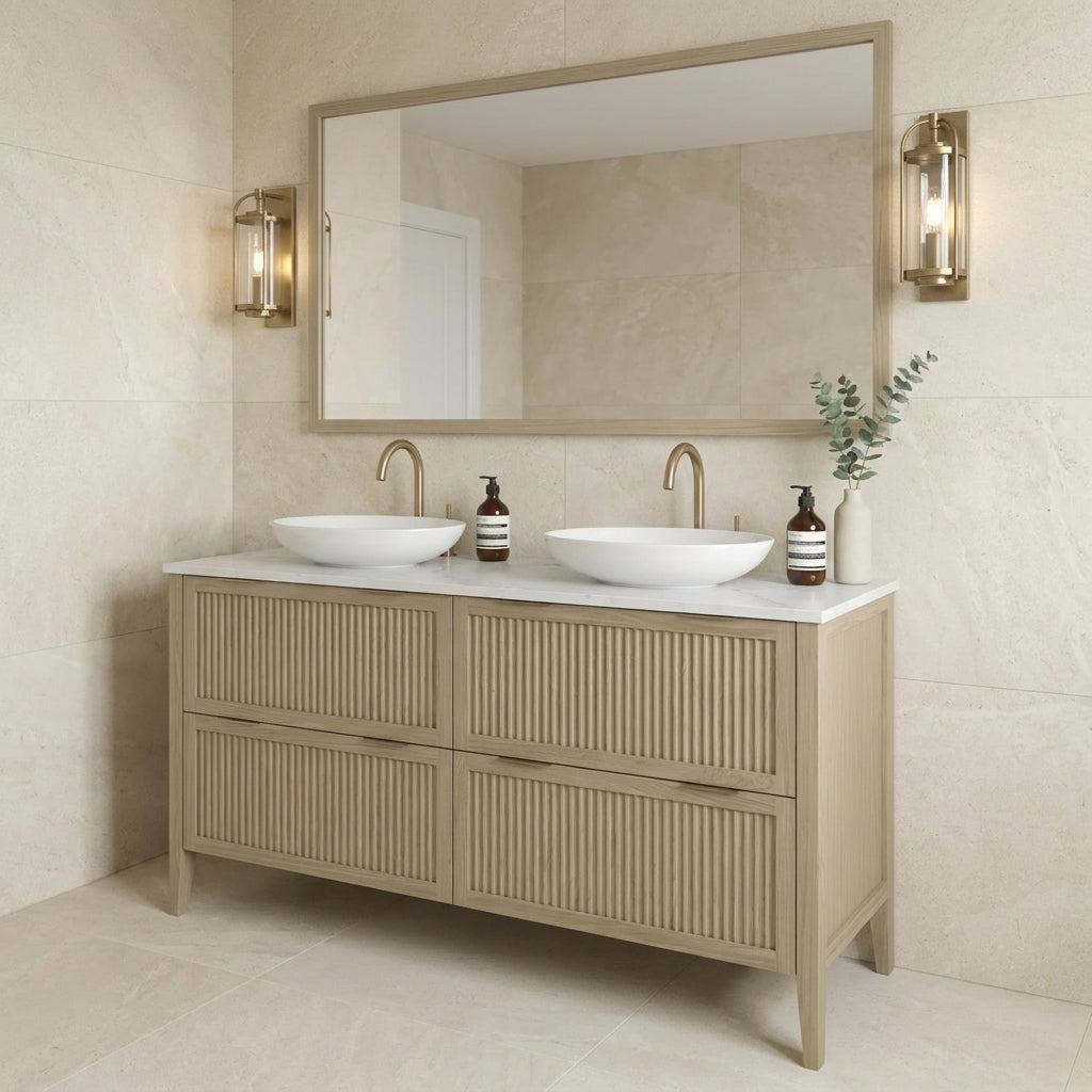 Mockridge Design Clifton 1400mm floor-standing double vanity in macchiato stained oak with soft milky-coffee tone and visible grain figuring across vertical fluted drawer fronts set within shaker frame borders, Calacatta quartz worktop, twin oval ceramic sit-on basins and twin brushed brass deck-mounted vessel mixer taps, styled in a calm contemporary English bathroom with warm cream limestone walls and pale honed limestone floor lit by diffuse ambient daylight