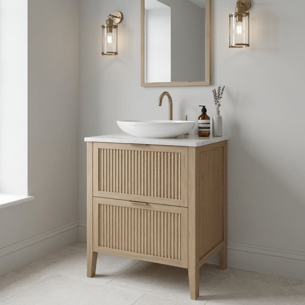 Mockridge Design Clifton 600mm floor-standing single vanity in smoke 5% stained oak with warm mid-pale tone softened by a defining cool smoky wash revealing oak grain figuring across vertical fluted drawer fronts set within shaker frame borders, Calacatta quartz worktop, oval ceramic sit-on basin and brushed brass deck-mounted vessel mixer tap, styled in a calm contemporary English bathroom with soft pale dove-grey walls and pale honed limestone floor